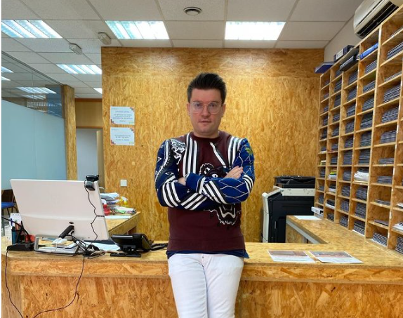 Antonio Barbeito posando delante de una mesa de atención al público, con pantalón blanco y jersey de rayas.